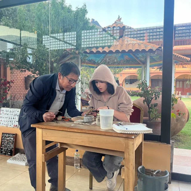 Two people focusing on a small object at a wooden table in a bright room with large windows overlooking a garden. A gas cylinder is nearby.