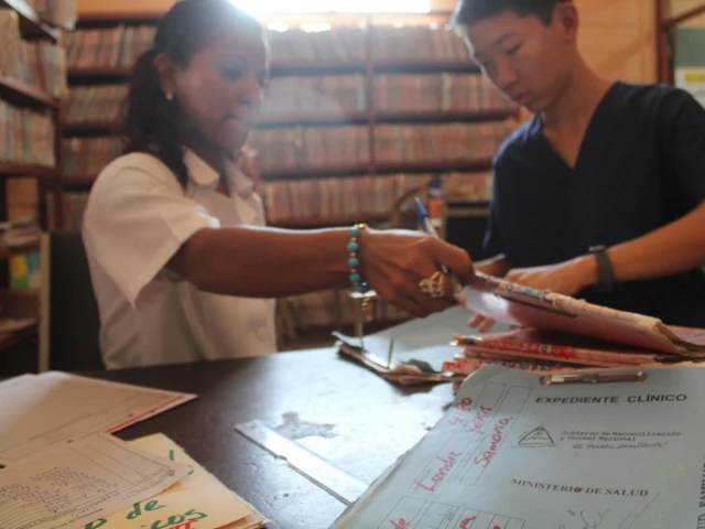 Two people organize folders and documents at a table in a room filled with shelves of files.
