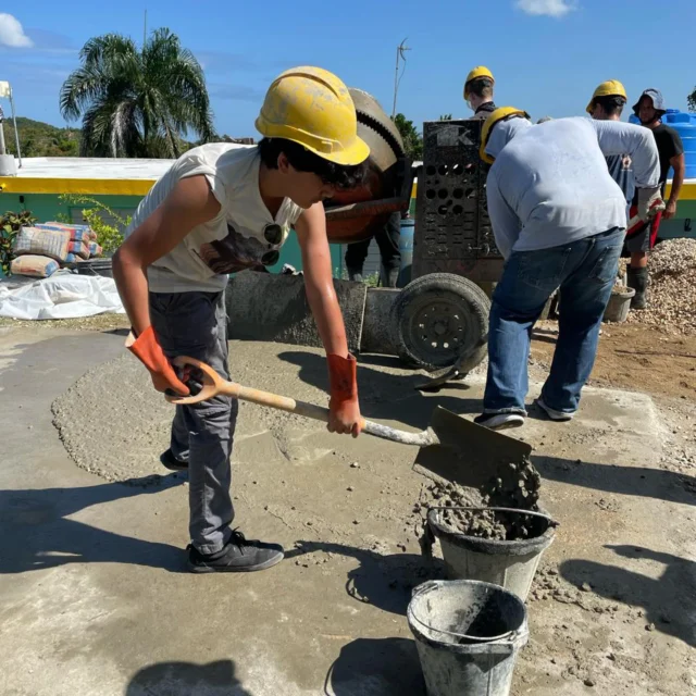 Workers in hard hats and gloves mix and pour concrete on a construction site under a clear blue sky.