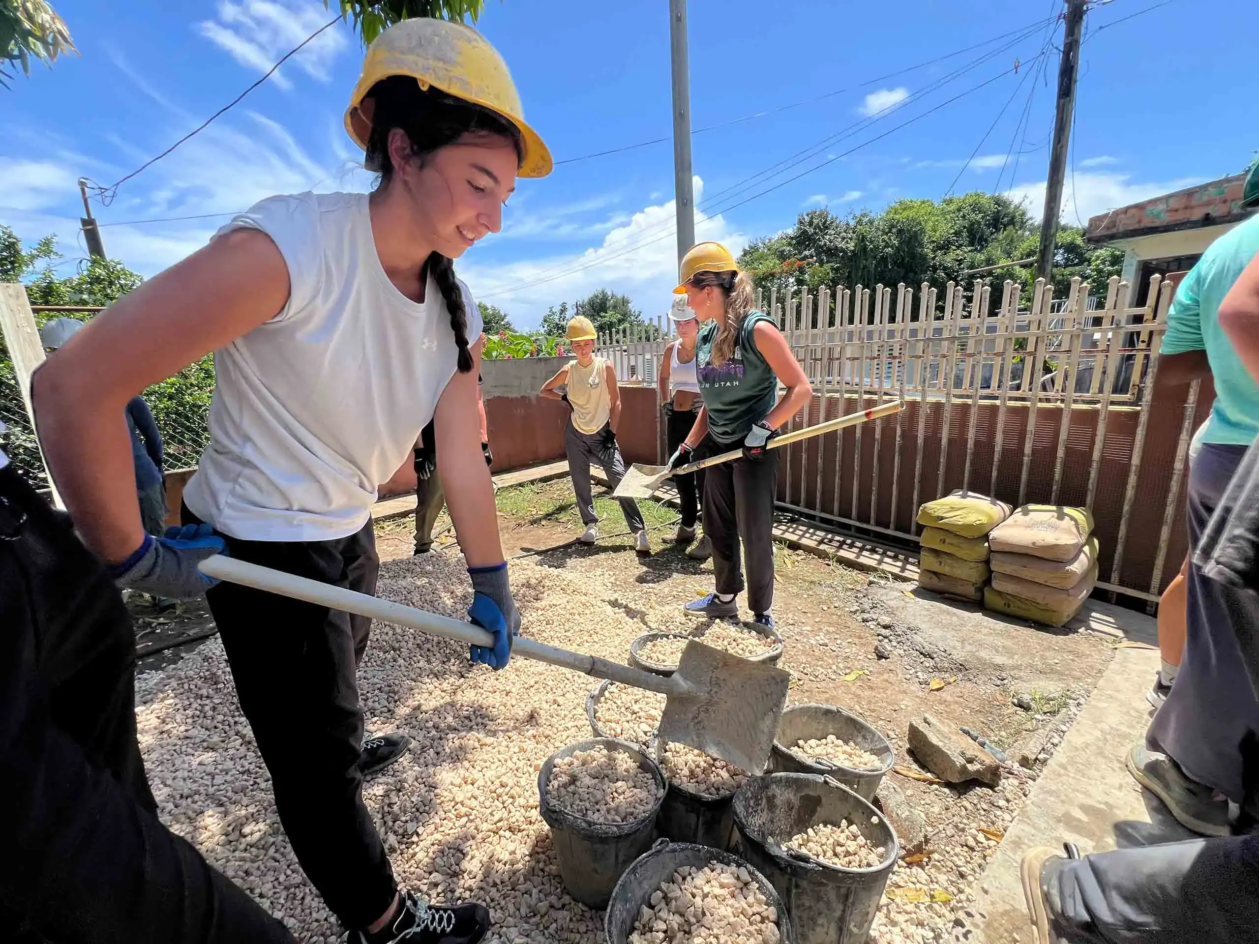 Several people on a Puerto Rico student service trip, wearing helmets and gloves, are shoveling gravel into buckets outdoors on a sunny day, next to a fence and bags of materials.