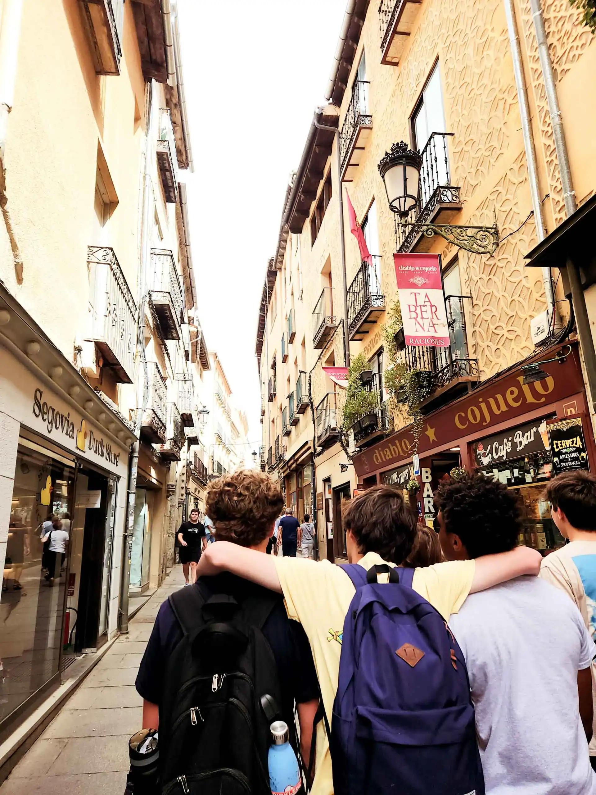 A group of young people with backpacks walk down a narrow street lined with shops and balconies during a school trip in Spain.