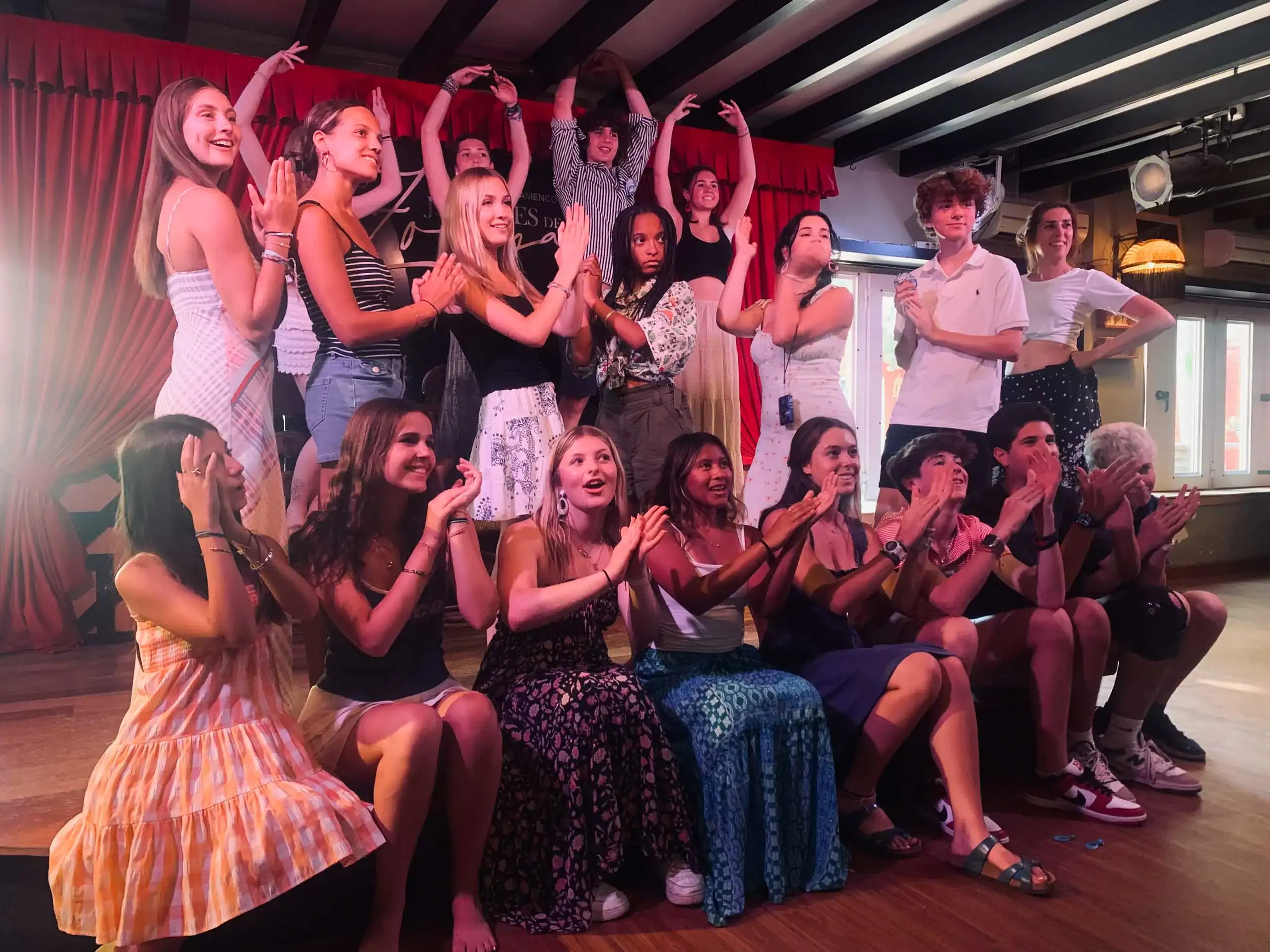 A group of young people pose and clap together on a stage, some standing and some sitting, with red curtains and spotlights in the background—capturing the excitement of their Spain school trip.