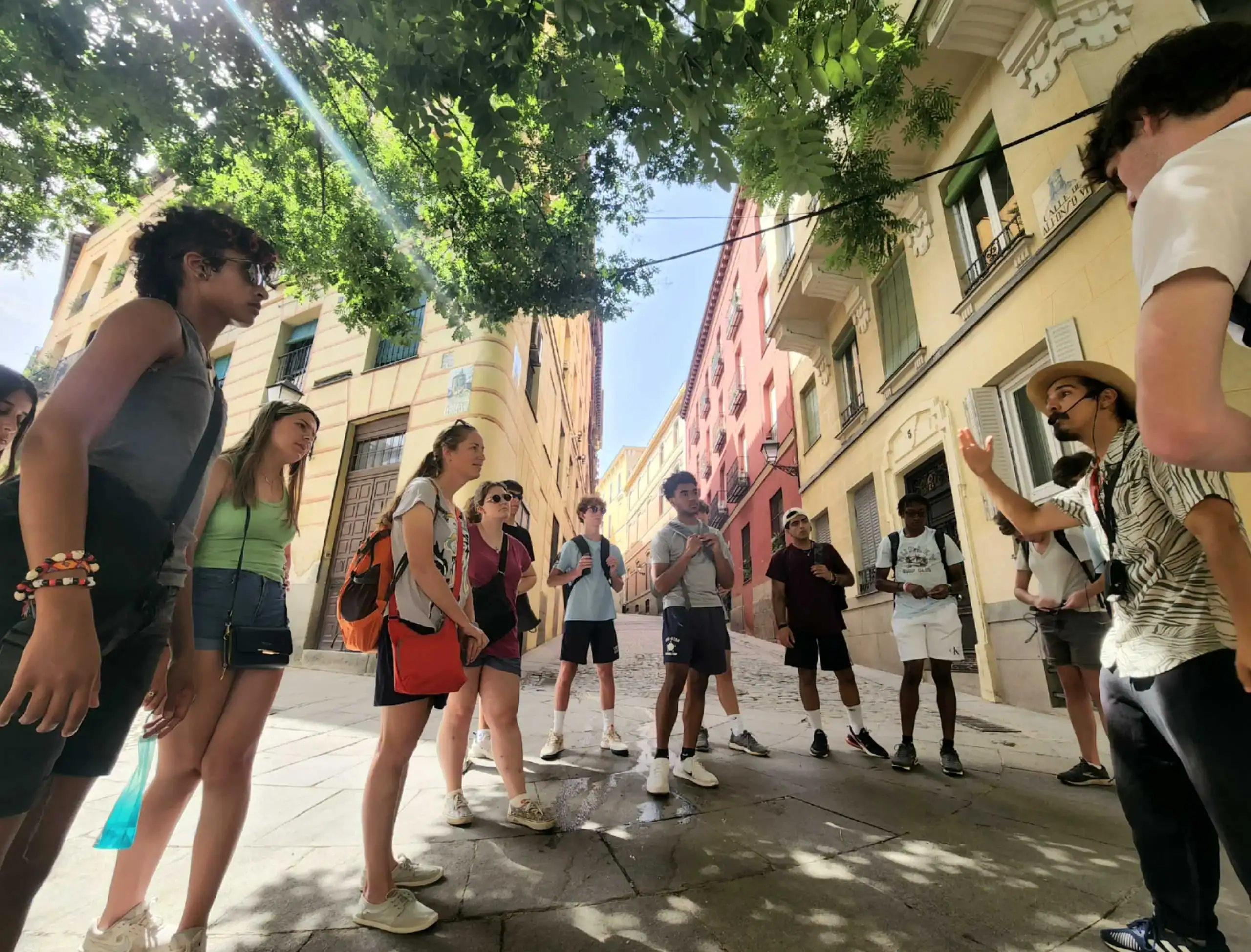 A group of students stand in a circle on a sunny street lined with colorful buildings, listening to a guide under the shade of green trees during their Spain school trip.