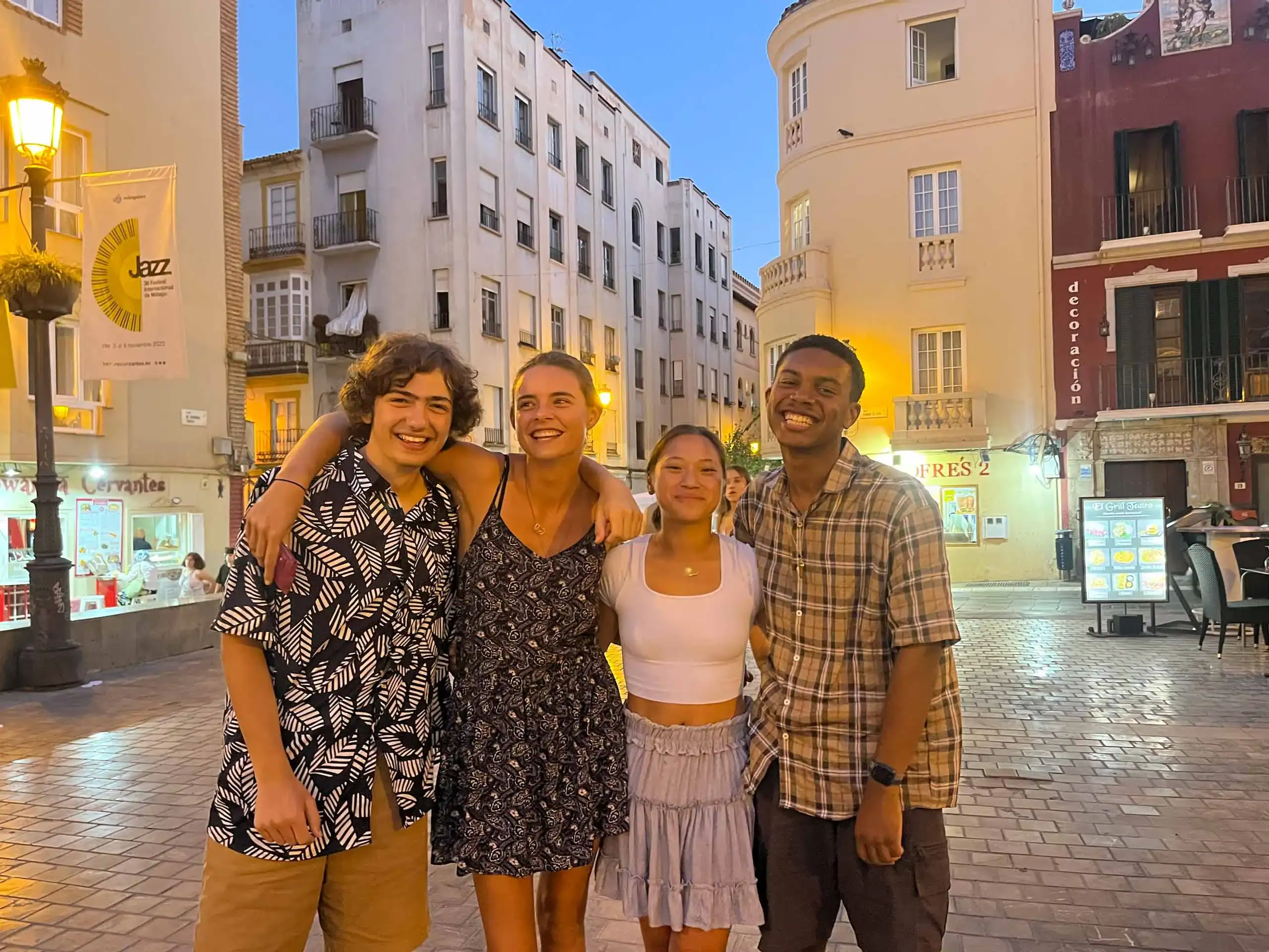 Four young adults stand together smiling in an outdoor plaza at dusk, enjoying their language immersion trip with buildings and streetlights glowing in the background.