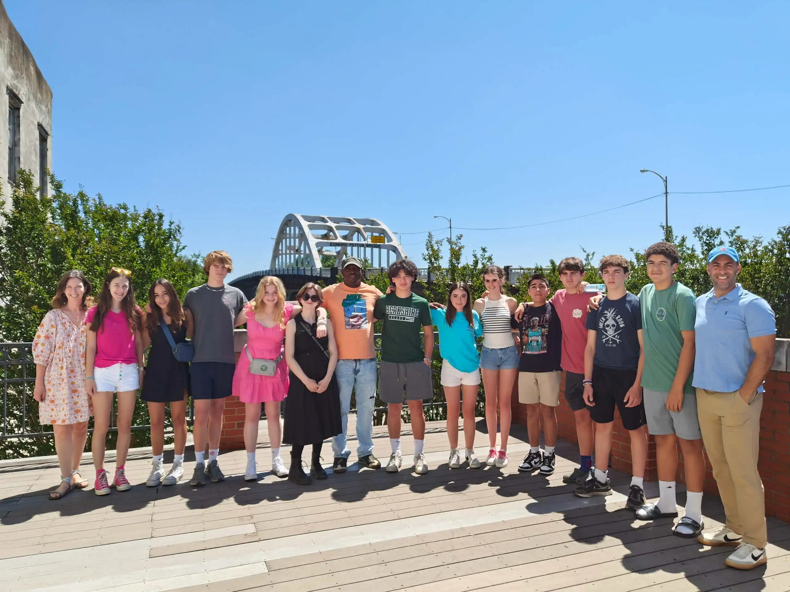 A USA school group travel outing to the Edmund Pettus Bridget with fifteen students standing side by side on a wooden deck outdoors overlooking the bridge.