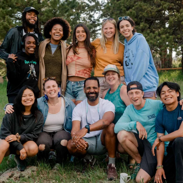 A diverse group of twelve people smiling and posing outdoors on a grassy field with trees in the background.