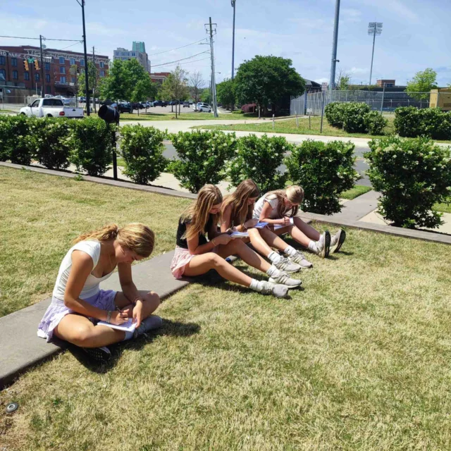 Five girls sit on the grass outside on a sunny day, each writing or drawing in notebooks, as part of a travel writing program, with a road, bushes, and stadium lights in the background.