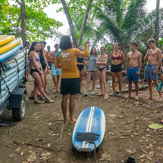 A group of people stand in a circle on a beach, listening to an instructor near a stack of surfboards and a single board on the ground.