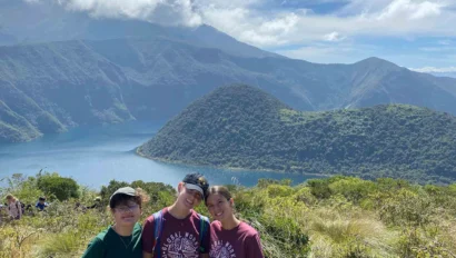 Three people stand on a grassy hill with a scenic view of a lake, green hills, and a cloudy sky in the background.
