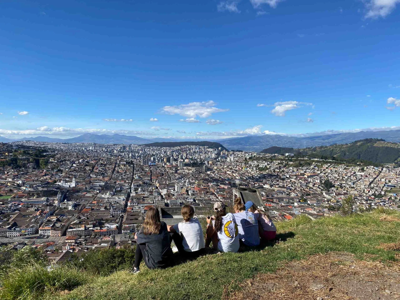 Five people sit on a hillside overlooking a sprawling cityscape under a clear blue sky.