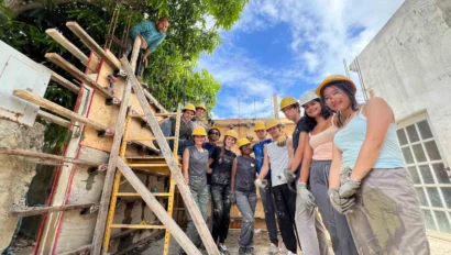 A group of people wearing hard hats and gloves stand next to a wooden structure and ladder at a construction site.