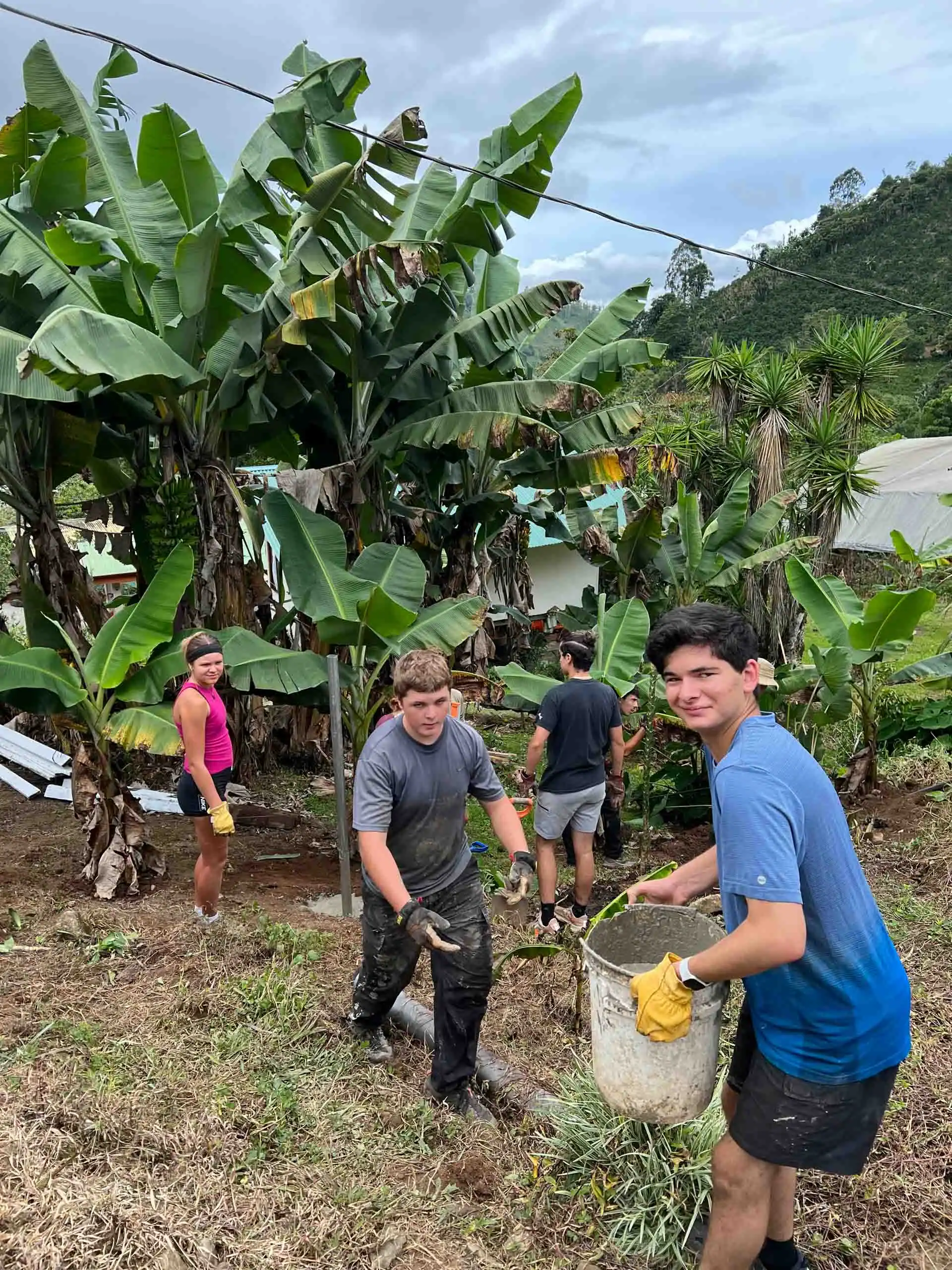 Four people work together outdoors, carrying a bucket and handling soil or plants near banana trees, with green hills and buildings in the background—a perfect snapshot of service trips for teens.