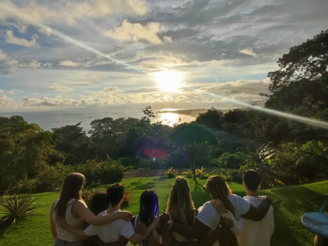 A group of people standing arm in arm on a grassy area, watching the sunset over a distant ocean.