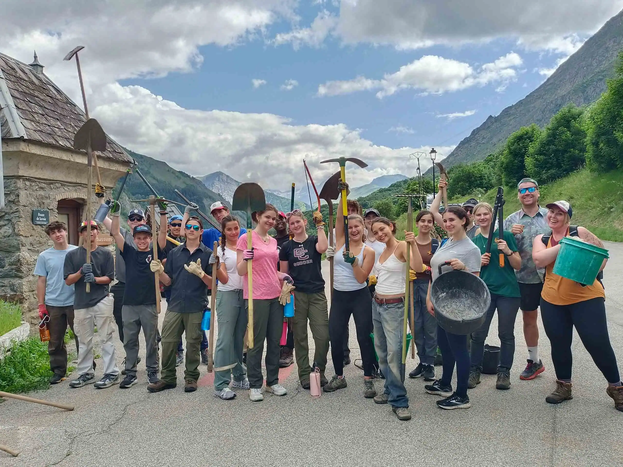 A group of people on a France student trip stand outdoors on a road in a mountainous area, holding various tools and buckets, posing for the camera.
