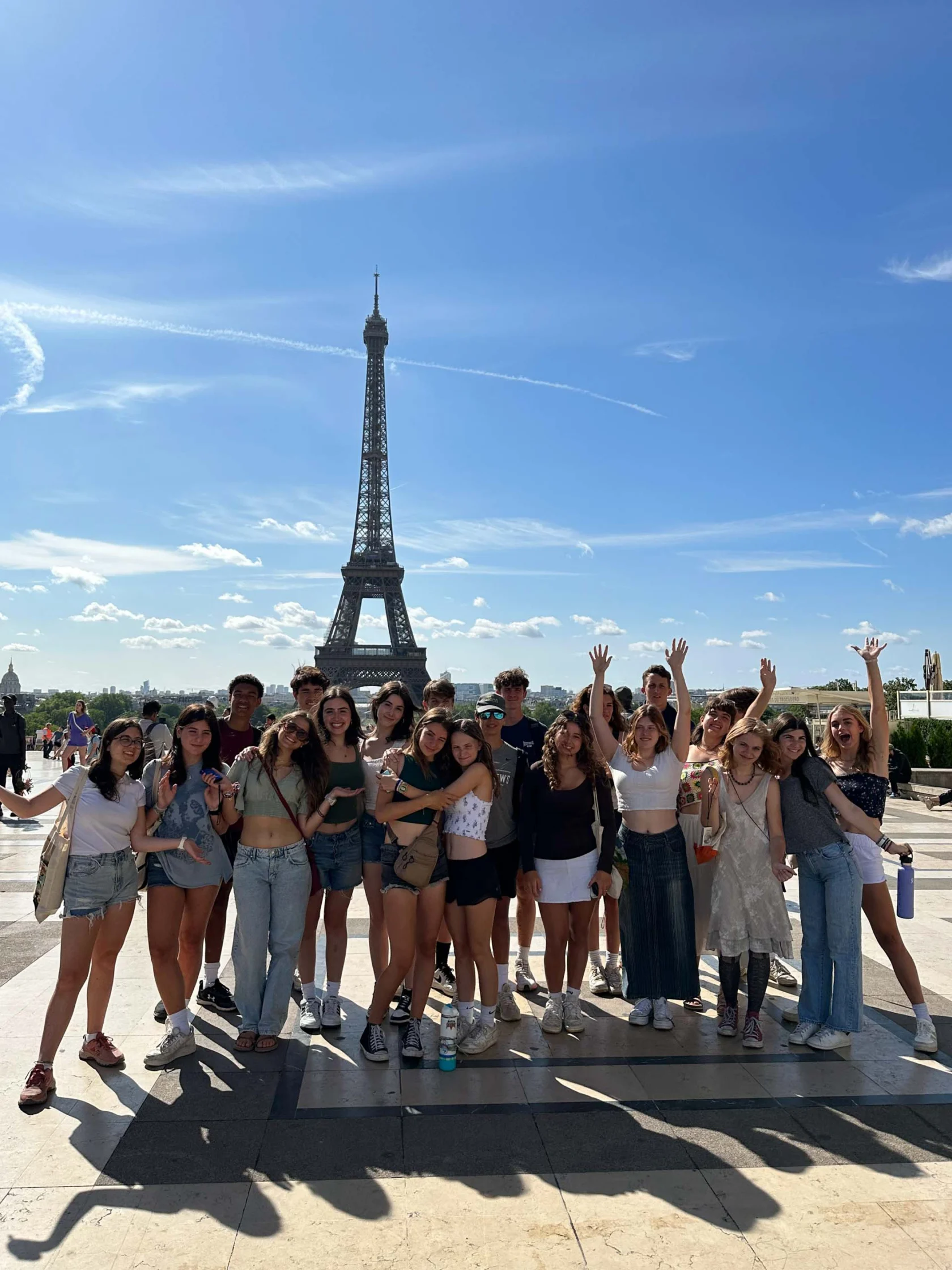 A group of people on a student trip to France poses and smiles in front of the Eiffel Tower on a sunny day with a clear blue sky.