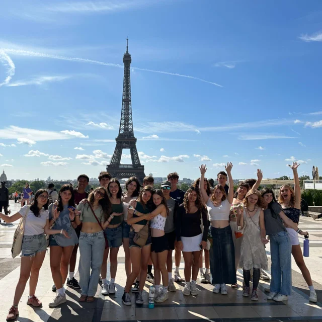 A group of people on a student trip to France poses and smiles in front of the Eiffel Tower on a sunny day with a clear blue sky.
