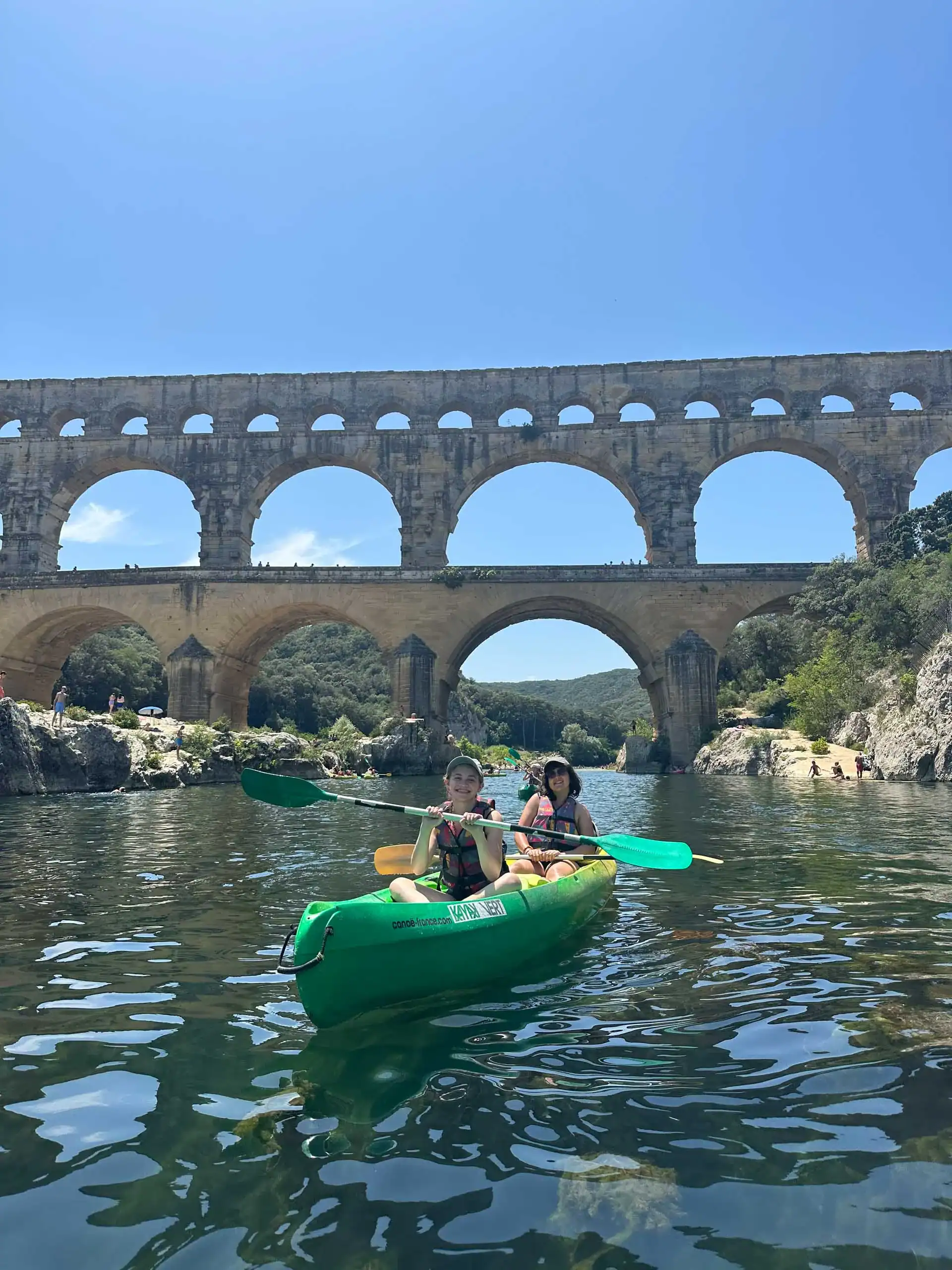 Two people kayaking on a calm river with the Pont du Gard Roman aqueduct in the background under a clear blue sky—a perfect scene from a student trip to France.