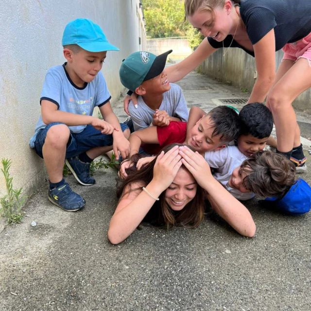 A group of kids and a young woman are playing on the ground outside, with the kids piled on top of the woman—perhaps reminiscing during a joyful student trip to France—as she lies on the pavement, smiling.