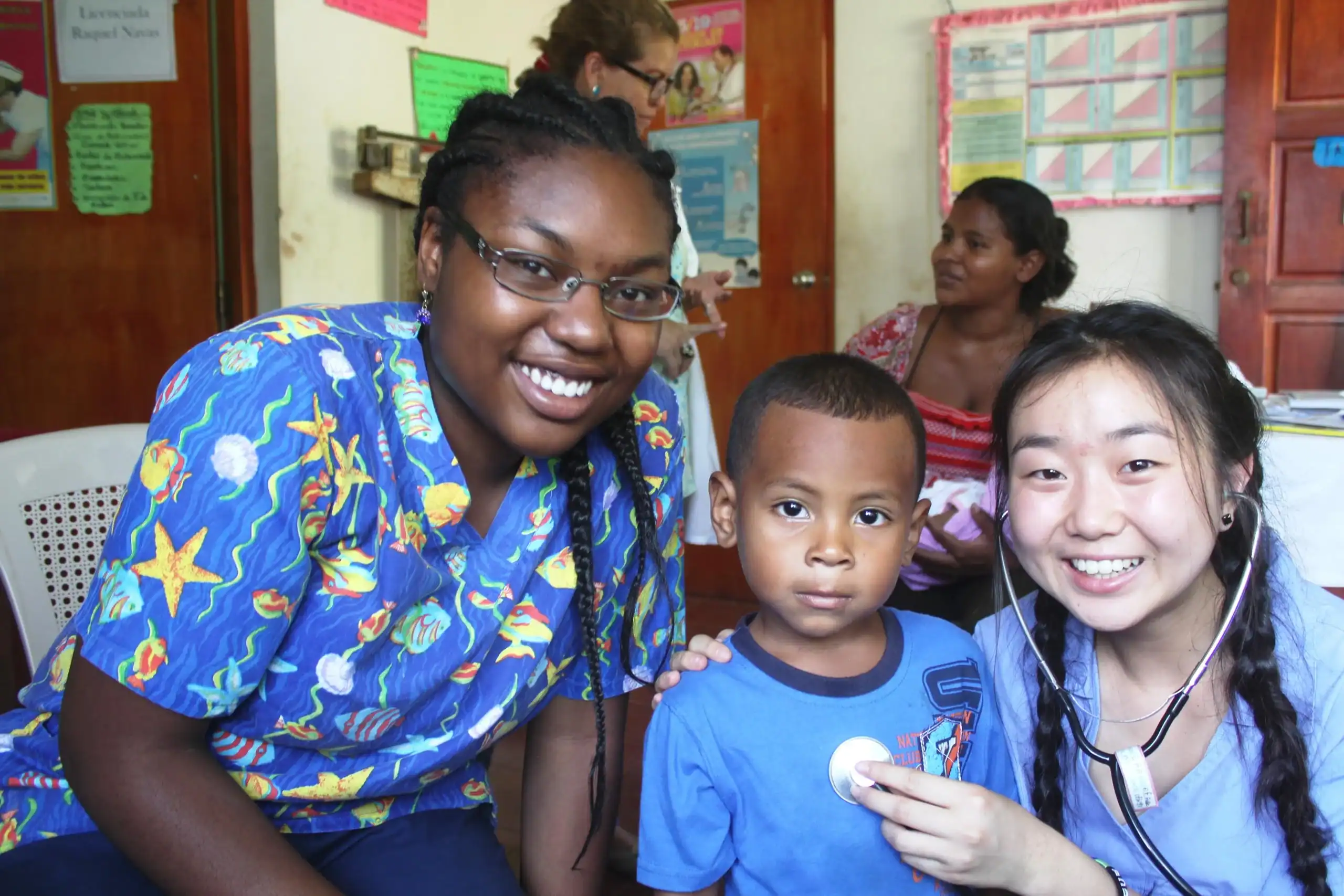 Two women and a young boy pose and smile in a brightly lit clinic room, likely part of a medical study abroad program; one woman places a sticker on the boy’s shirt while others are seen in the background.