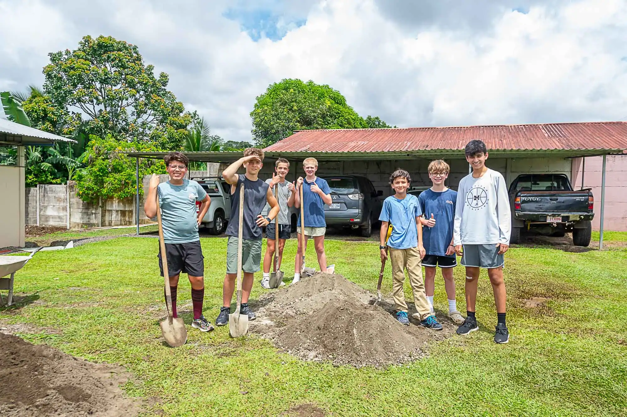 Seven boys with shovels, part of a middle school travel program, stand around a pile of dirt on a grassy lawn, with cars and a shed in the background on a partly cloudy day.