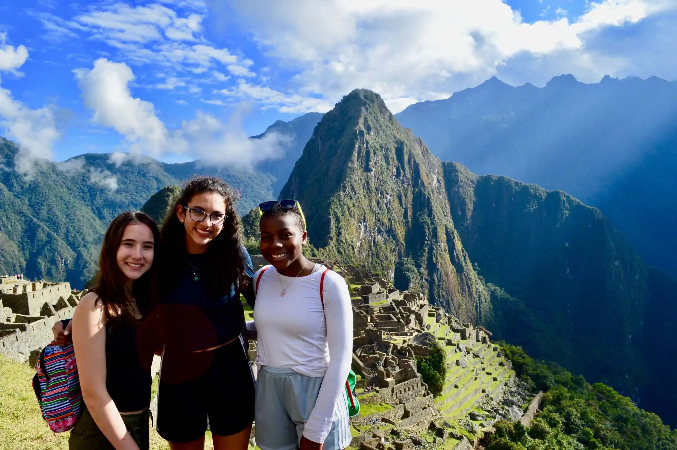 On a Peru student trip you see three people smiling at Machu Picchu, with mountains and ruins in the background.