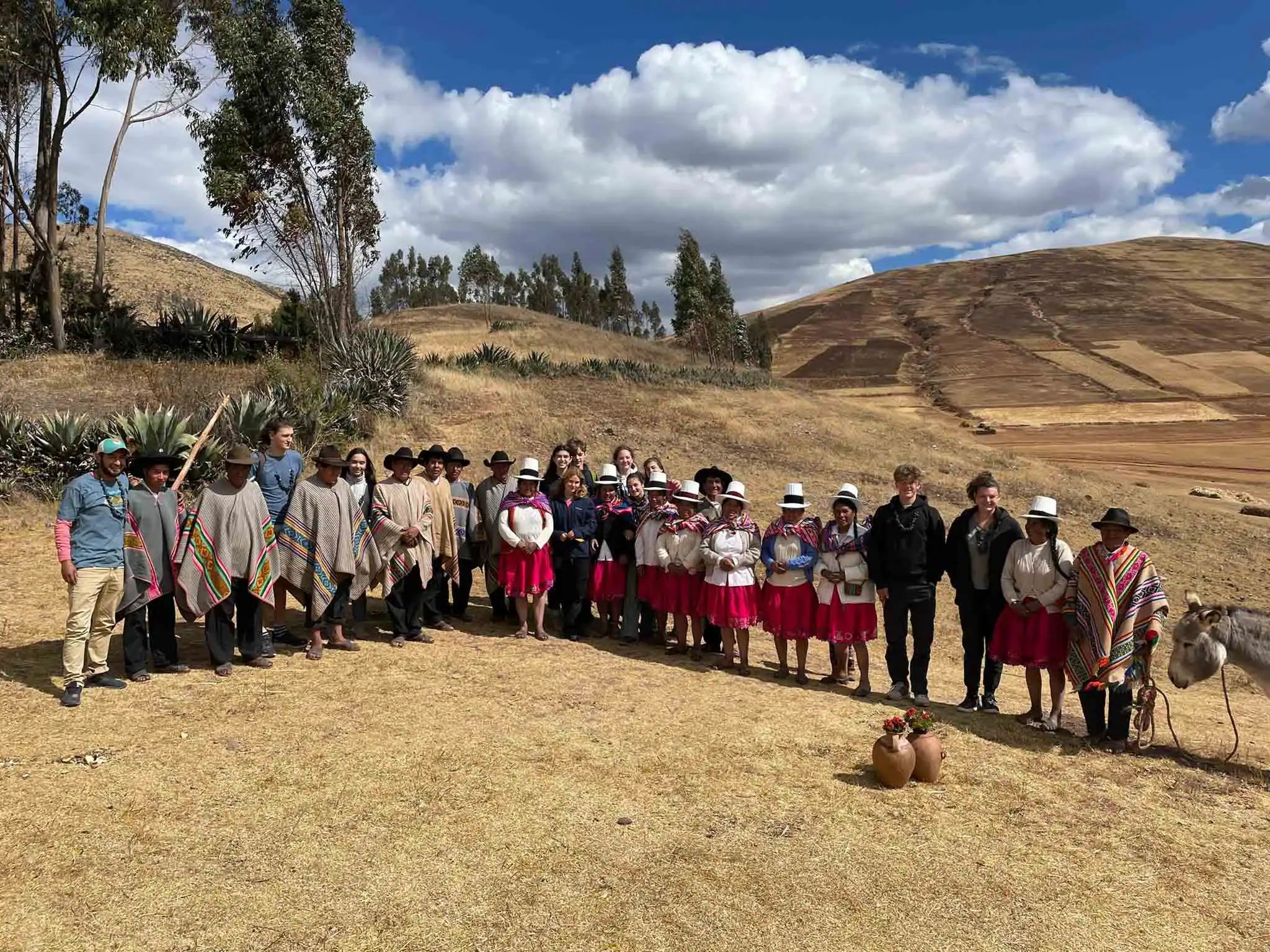 A group of people on a Peru student trip are wearing traditional clothing stands in a grassy field with hills and trees in the background on a sunny day.