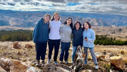 Five people standing together on a rocky hill with a mountainous landscape in the background under a cloudy sky.
