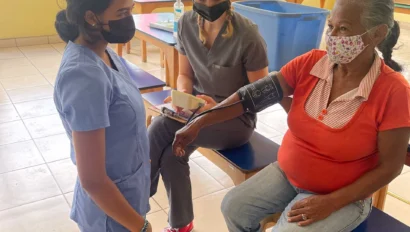 Two healthcare workers check a seated elderly woman's blood pressure in a bright room.