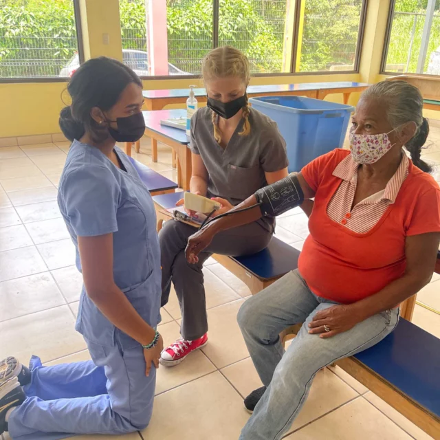 Two healthcare workers check a seated elderly woman's blood pressure in a bright room.
