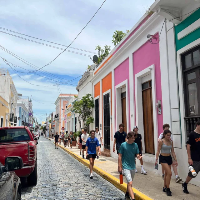 People walk along a colorful street with vibrant buildings in a historic district. Cars are parked along the cobblestone road.