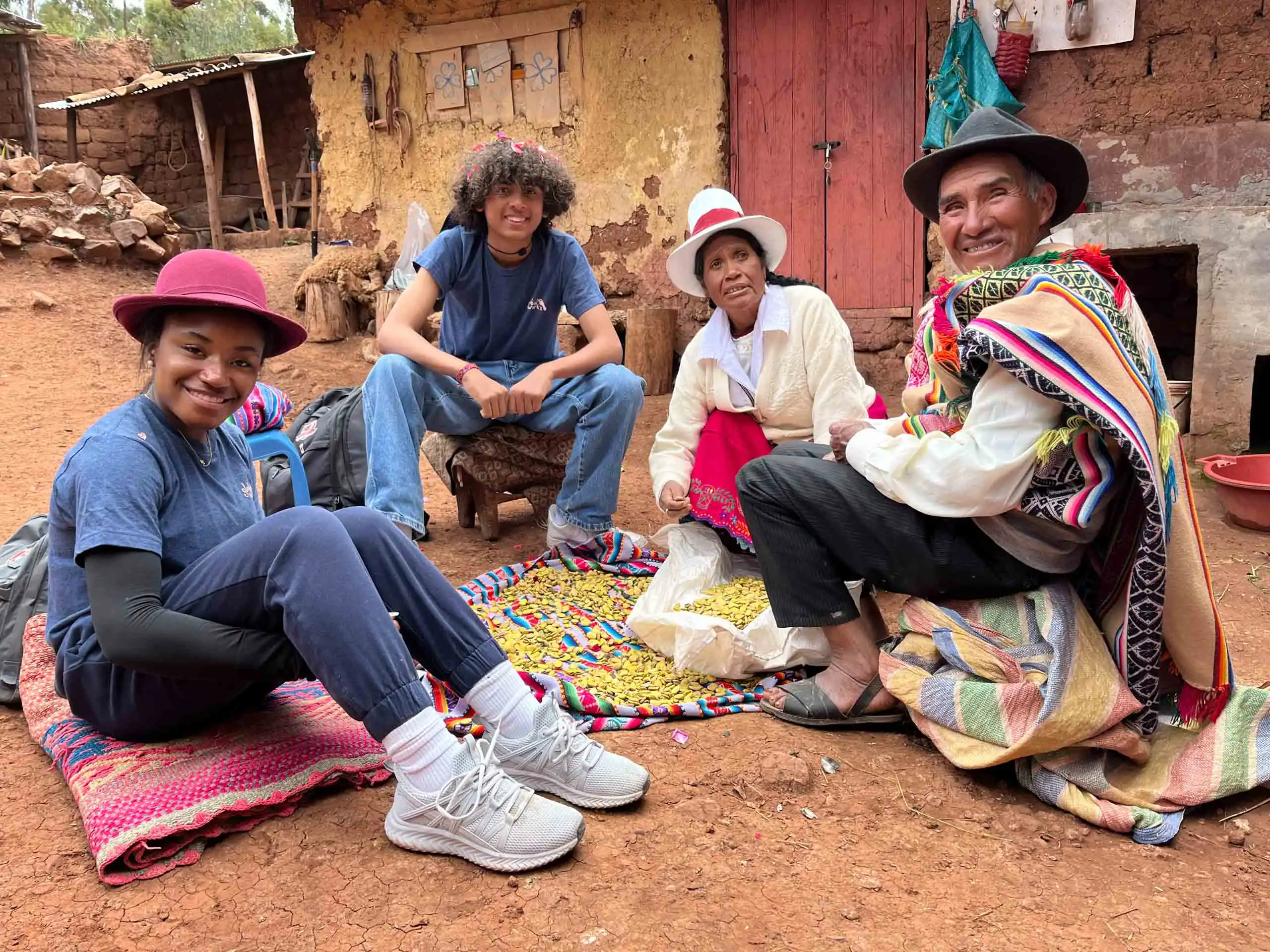 Four students on a teen tour in a rural setting are seated on a blanket, surrounded by colorful textiles and corn. They are wearing casual and traditional clothing, with a rustic building in the background.