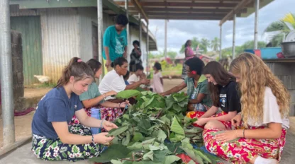 People sitting on the ground, weaving leaves together, surrounded by a colorful mat under a covered outdoor area.