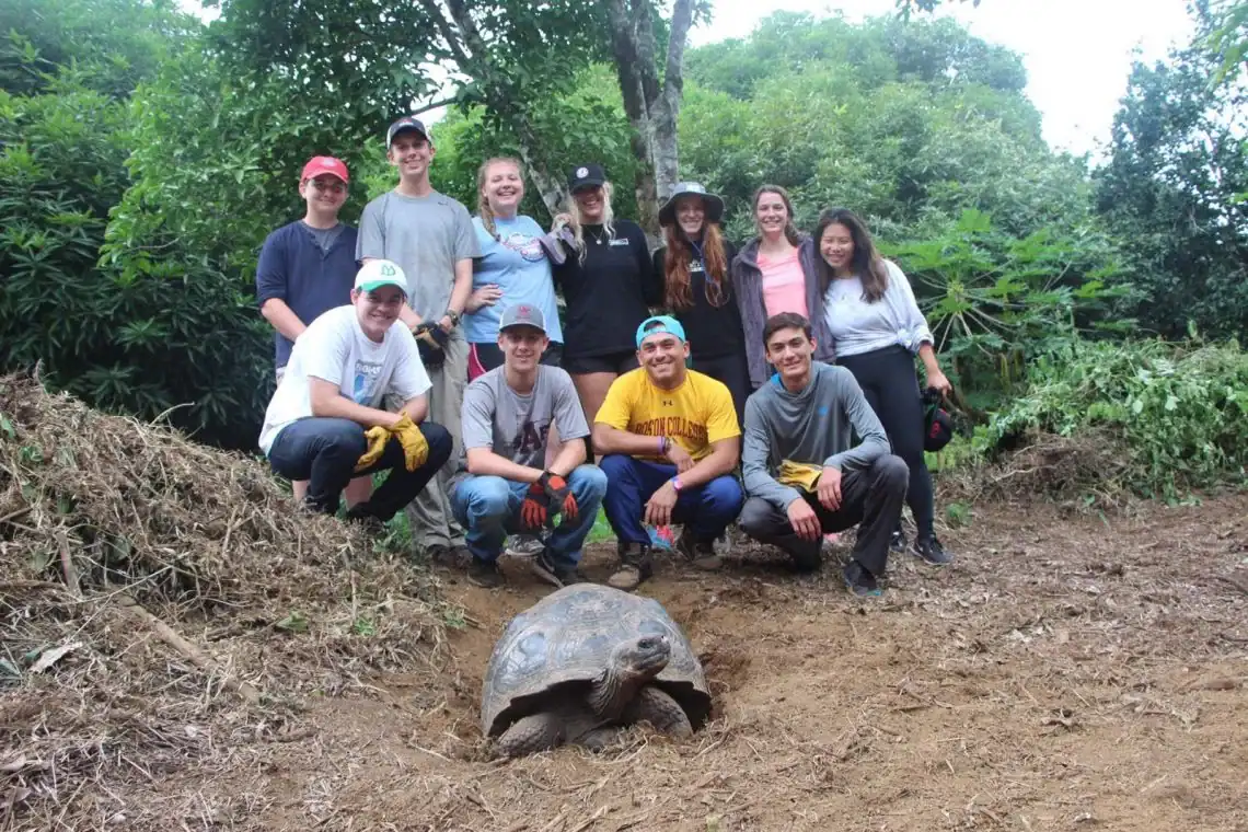 A group of ten people wearing casual clothes stands behind a large tortoise in a lush, green outdoor setting on an immersive teen tour experience.