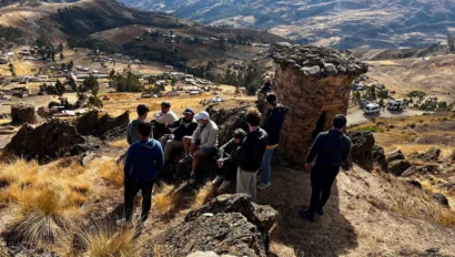 A group of people gather on a rocky hill with a scenic view of a village and mountains in the background.