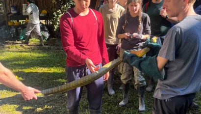 A group of people stands outdoors examining a large snake held by two individuals. They're surrounded by greenery and wearing casual outdoor attire.