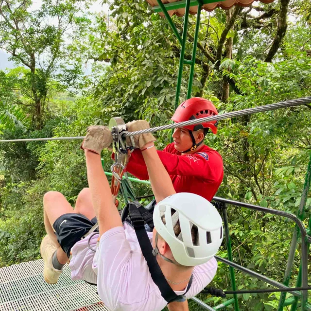 Person in white helmet preparing for a zipline ride with the assistance of an instructor in a red helmet, surrounded by lush green forest.