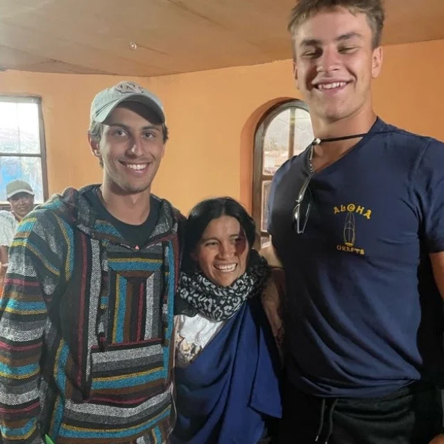 Three people stand indoors smiling at the camera; two young men flank an older woman. The warm-colored walls and natural light evoke the relaxed mood of summer travel in this casual setting.