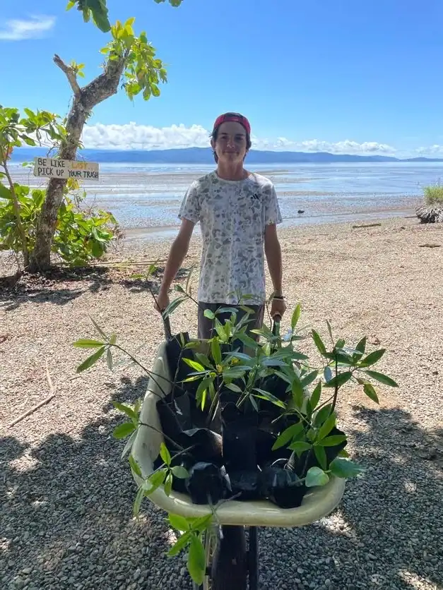 A person stands on a beach, pushing a wheelbarrow full of young plants during a conservation trip. In the background, a sign reads, "Be like. Pick up your trash".