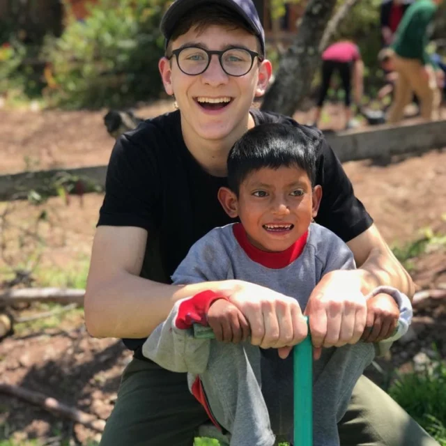 A smiling young adult and a child sit on a seesaw in an outdoor setting with trees and people in the background.
