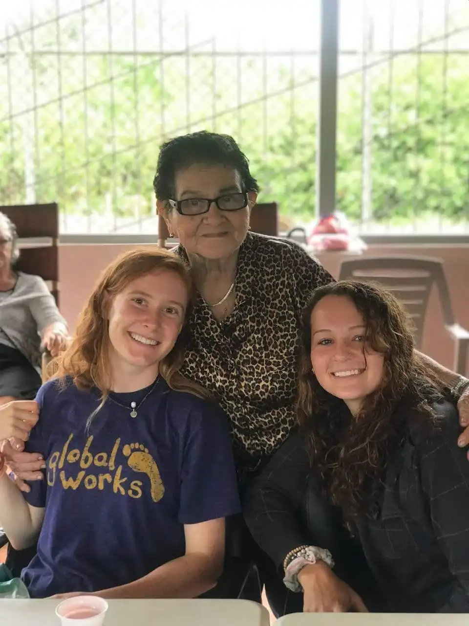 Three women pose together and smile at a table outdoors. The older woman stands behind two younger women, one in a “global works” t-shirt while experiencing their student travel to Costa Rica. Greenery is visible in the background.