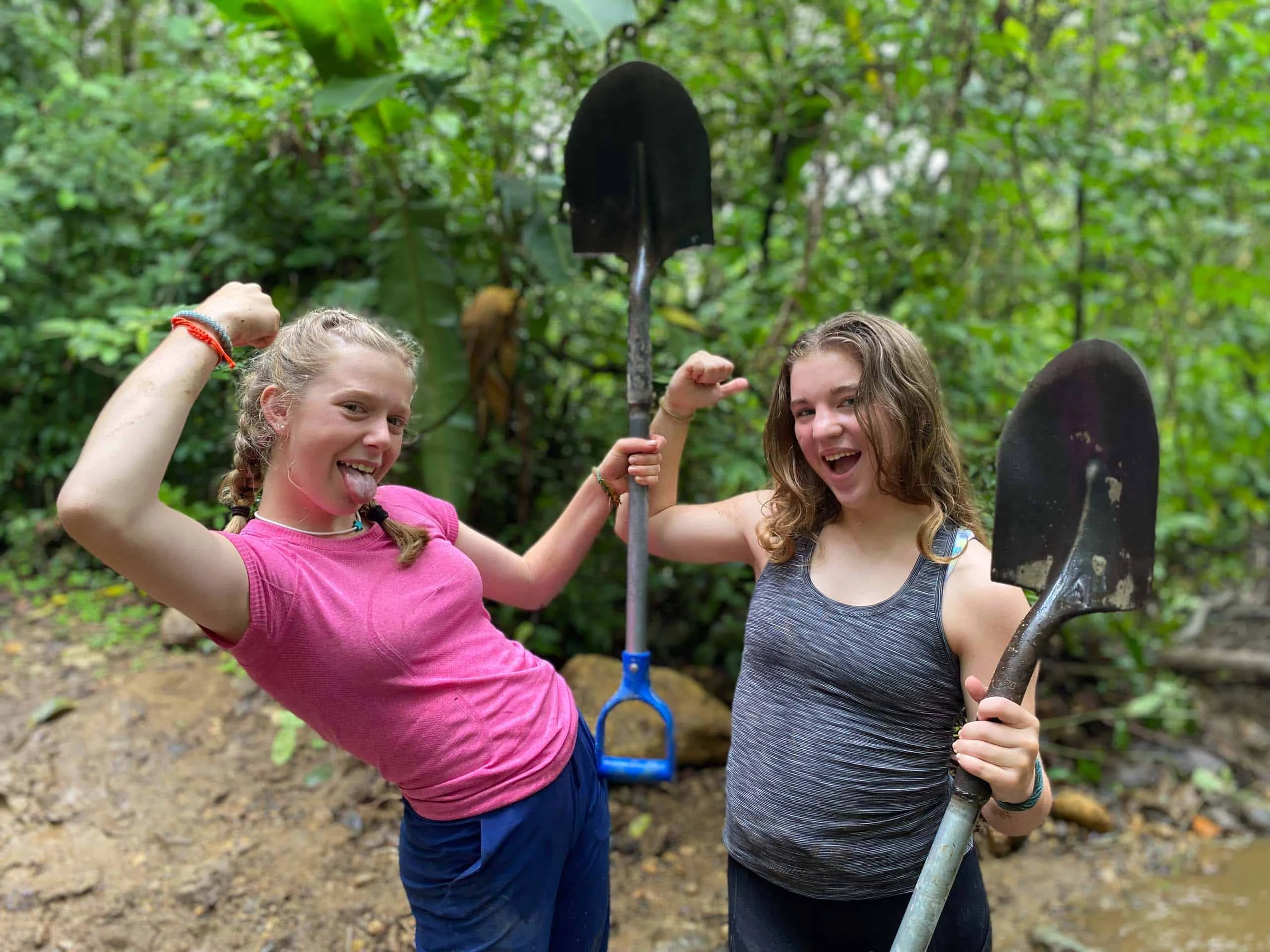 Two young women, part of a Global Works student travel program, pose and flex their arms while holding shovels in a muddy, forested outdoor setting.