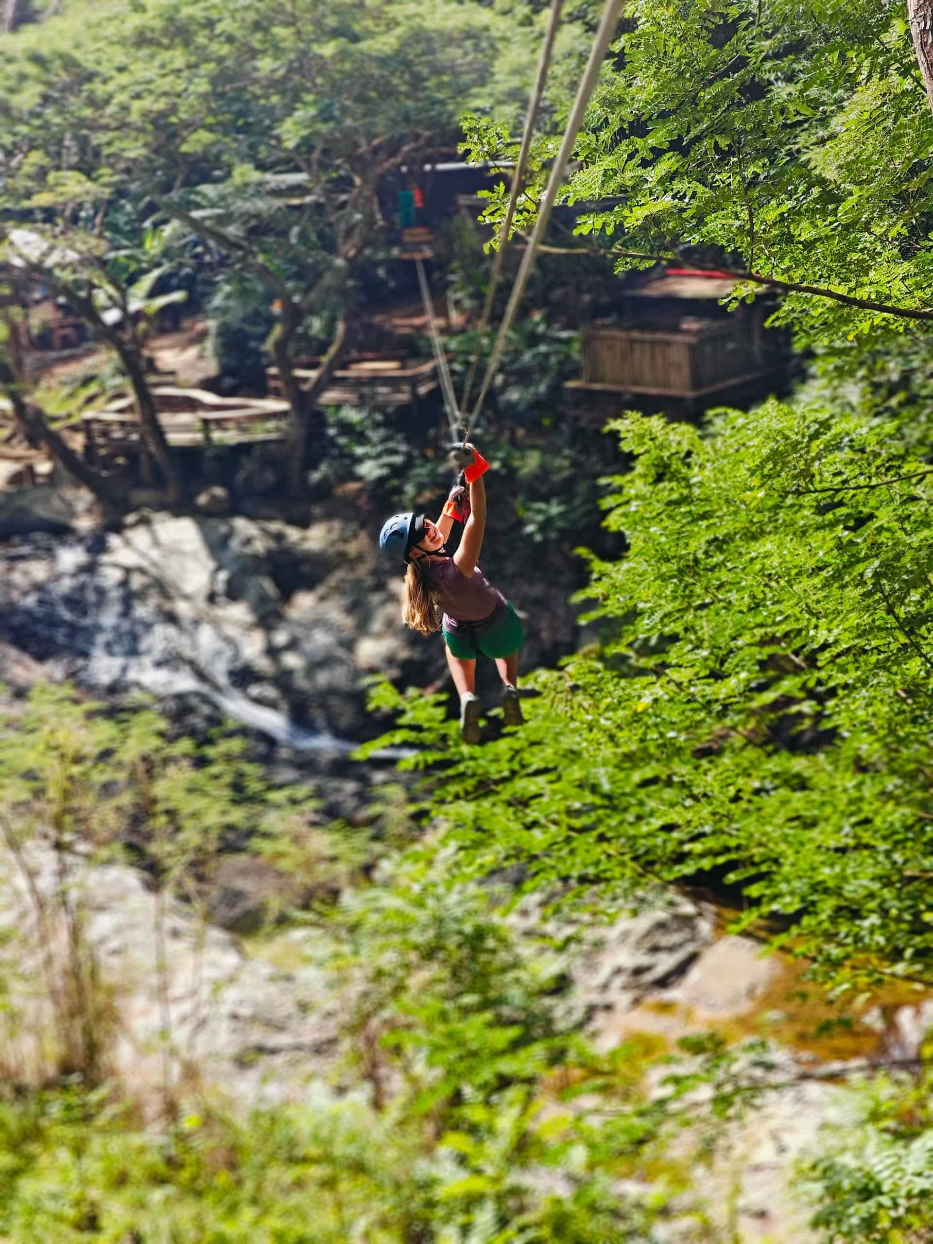 A person wearing a helmet and harness glides on a zip line above a forested area, making this the perfect adventure travel for teens with trees and a river below.