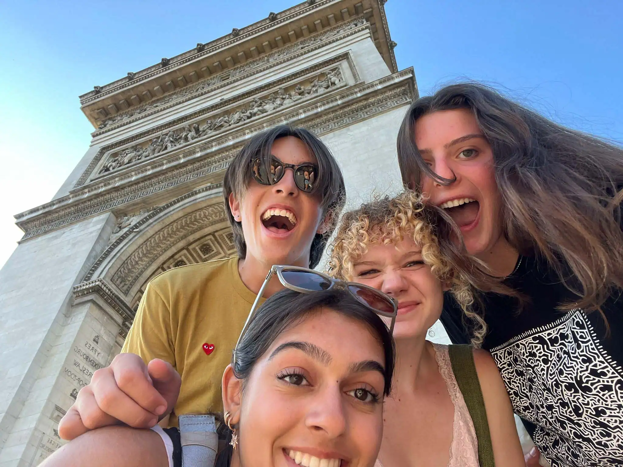 Four smiling young people pose for a selfie in front of the Arc de Triomphe on a sunny day, capturing memories from their French Immersion Teen Travel Program.