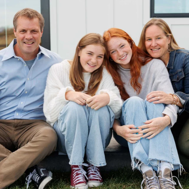 A family of four sits together on a porch step, smiling at the camera.