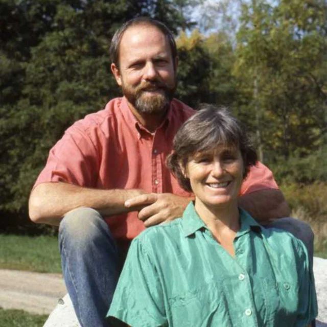 A man and woman pose outdoors on a sunny day, sitting on a rock with trees in the background. The man wears a red shirt, and the woman wears a green shirt.