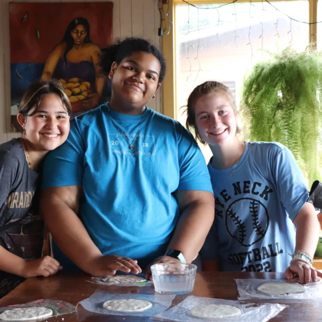 Three people smiling at a table with tortillas, a bowl of water, and plants in the background.