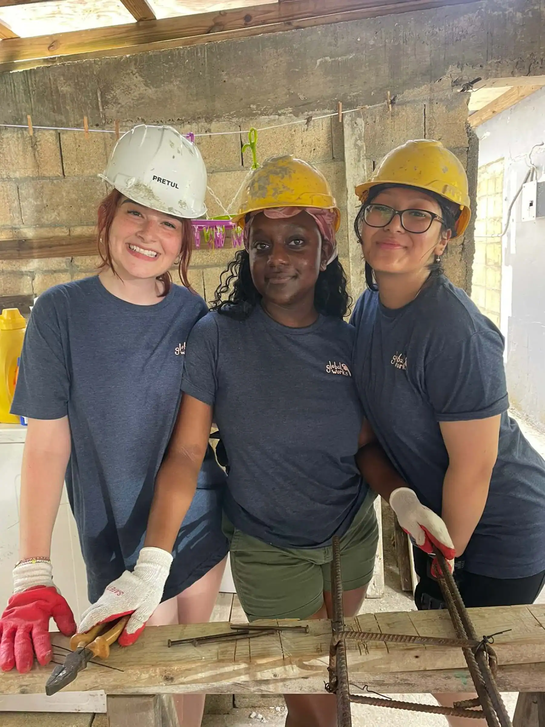 Three student travelers during a school group travel program wearing hard hats, gloves, and matching blue shirts as they pose indoors at a construction site, smiling while working with metal rods and wooden planks.