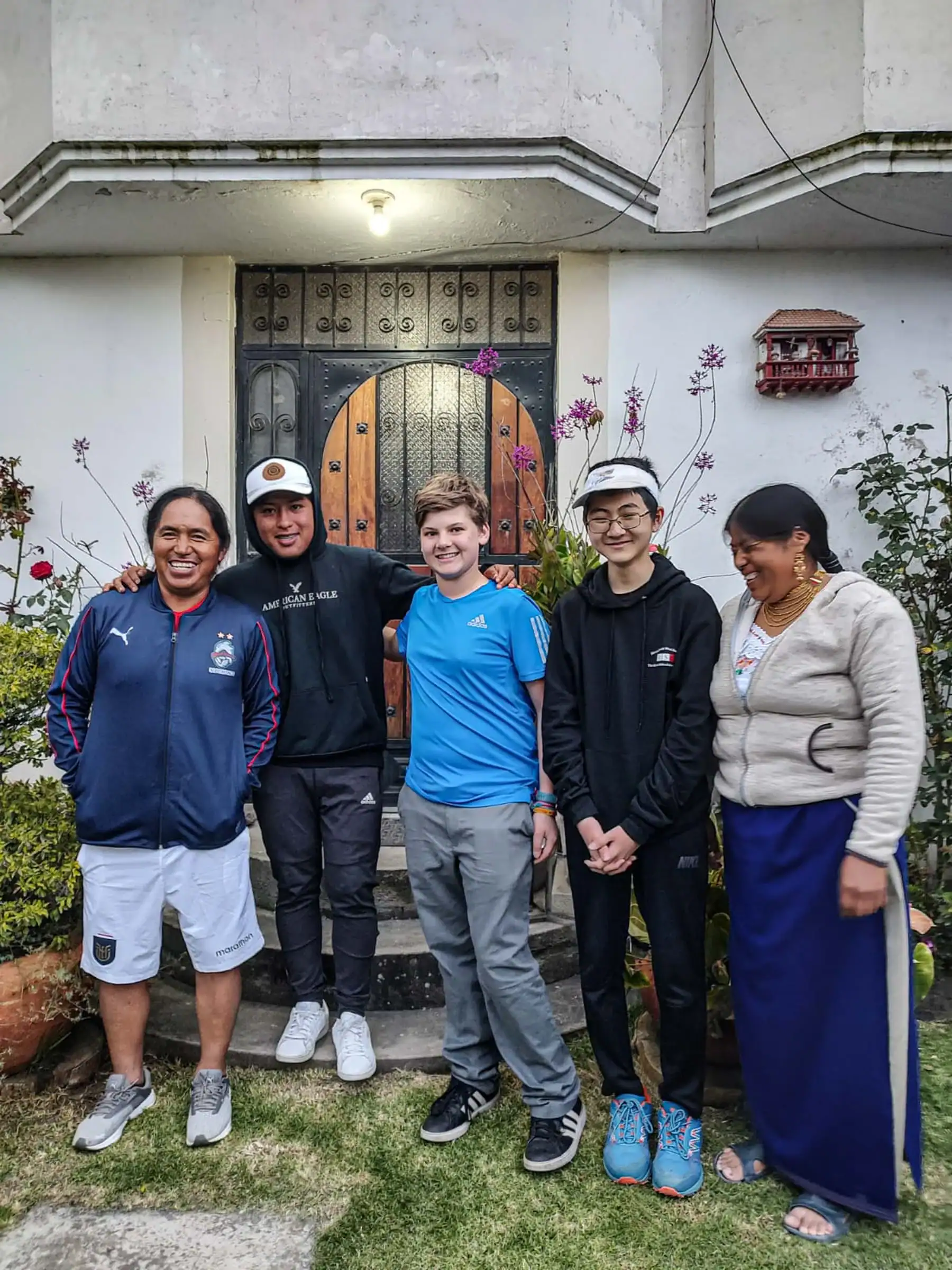 high school students stand and smile in front of a doorway framed by potted plants and flowers, sharing a moment together during their homestay on their Spanish immersion trip; one woman looks down and smiles while the others face the camera.