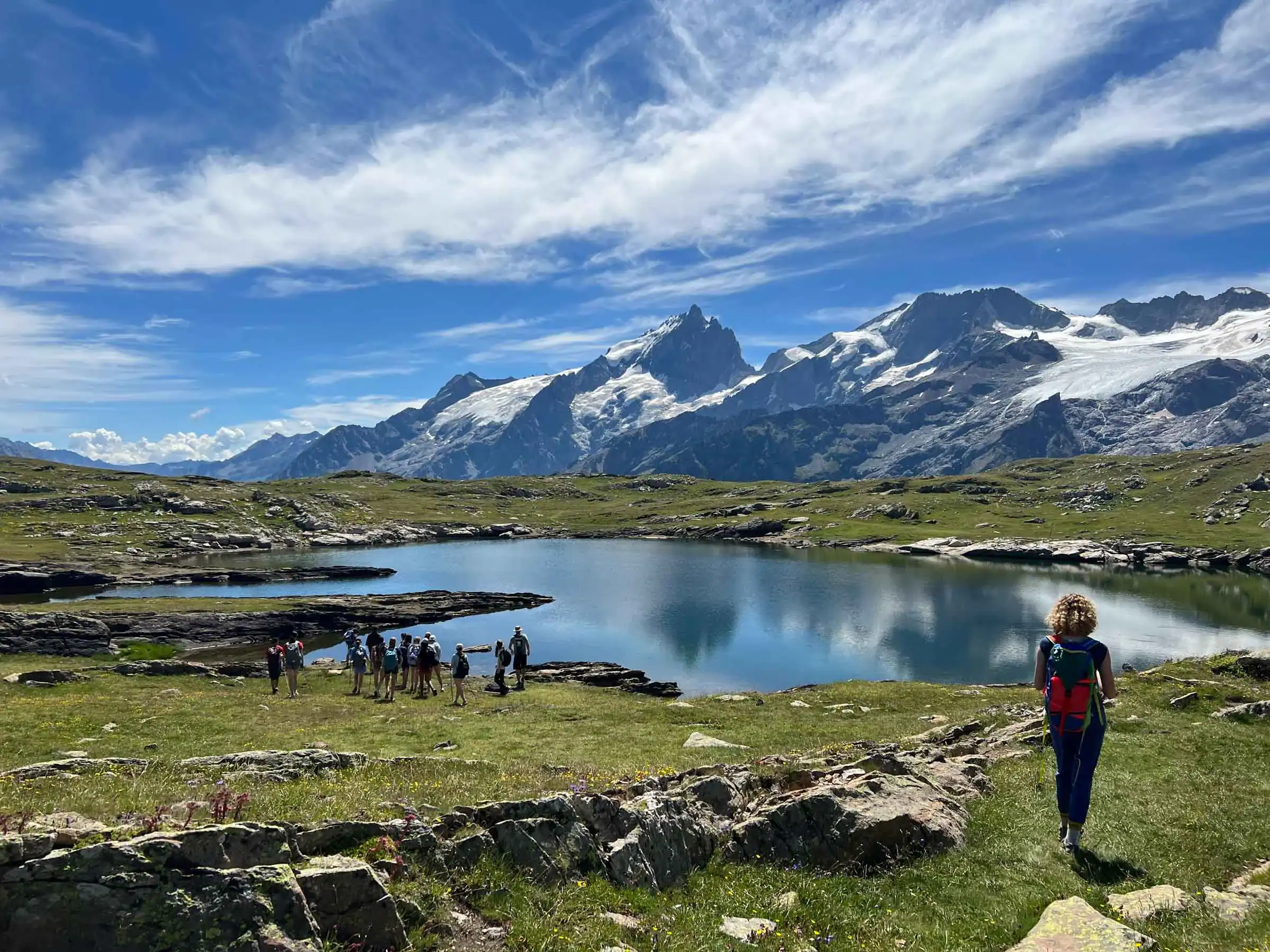 A group of student travelers by a lake with mountains in the background, while one person with a backpack—part of a student travel program—walks toward them under a partly cloudy sky.