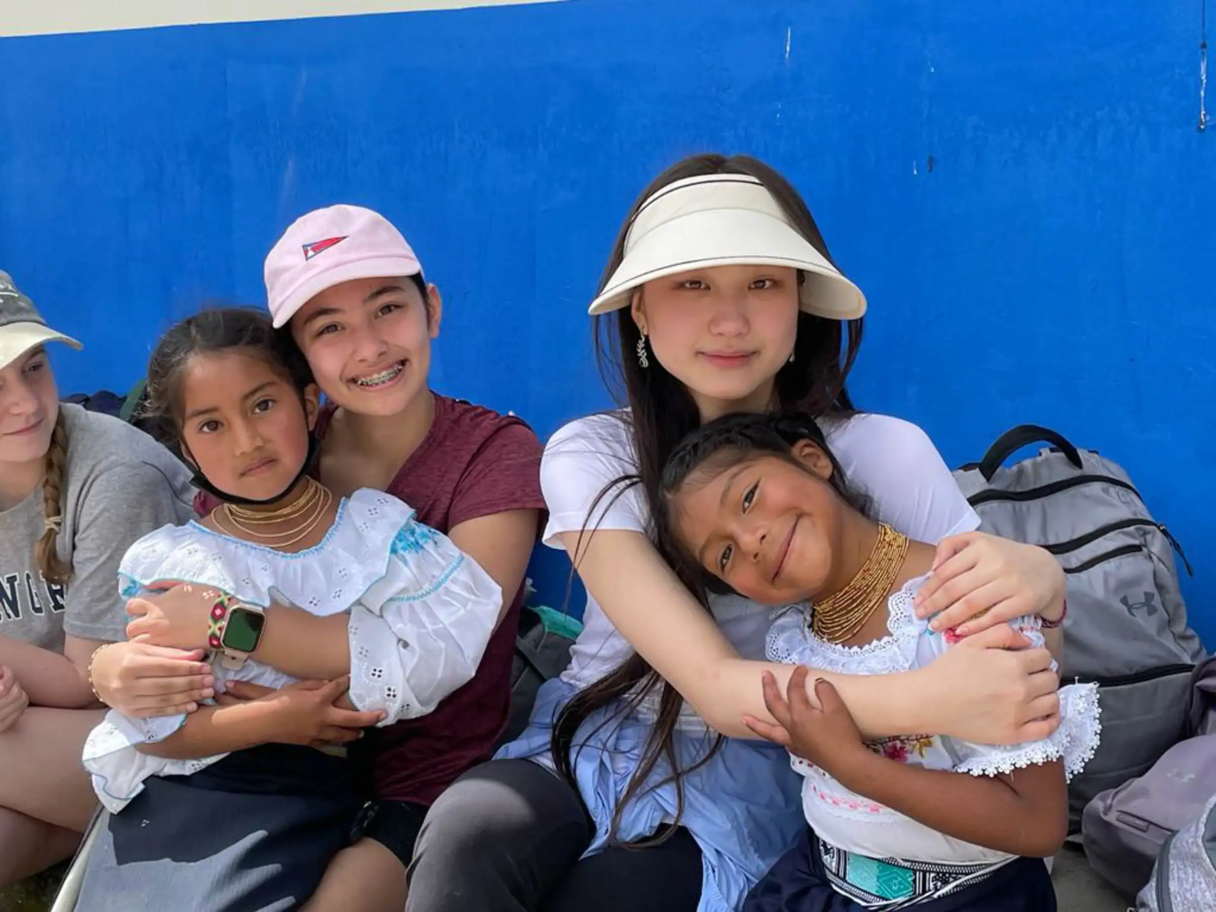 Three girls pose with two young children wearing traditional attire against a blue wall. One girl wears a visor, another a pink hat.
